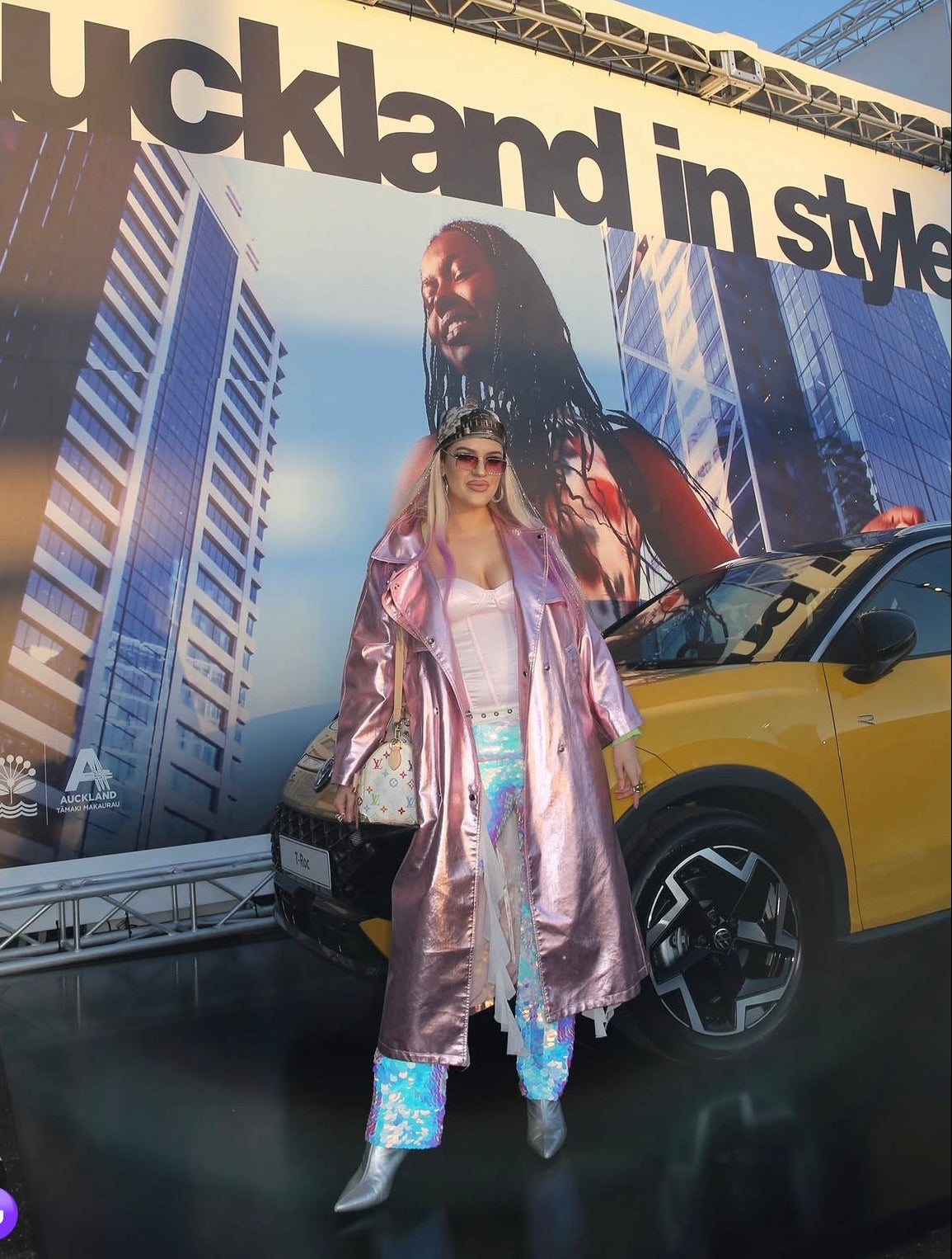 A woman stands in front of a large 'Auckland in style' event backdrop, AT NZ FASHION WEEK wearing a shiny metallic pink duster coat over a white outfit with NOVA SEQUIN FLARE TROUSERS and silver boots. A yellow car is partially visible beside her, and an Auckland Council Events logo appears on the backdrop.