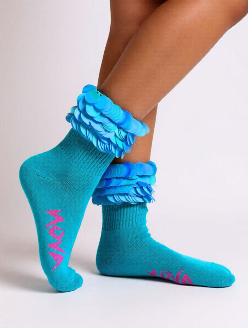 A pair of bright turquoise crew socks with layered ruffle cuffs in shades of blue and pink text reading 'nova' on the soles, shown on a model's feet against a light gray studio background.