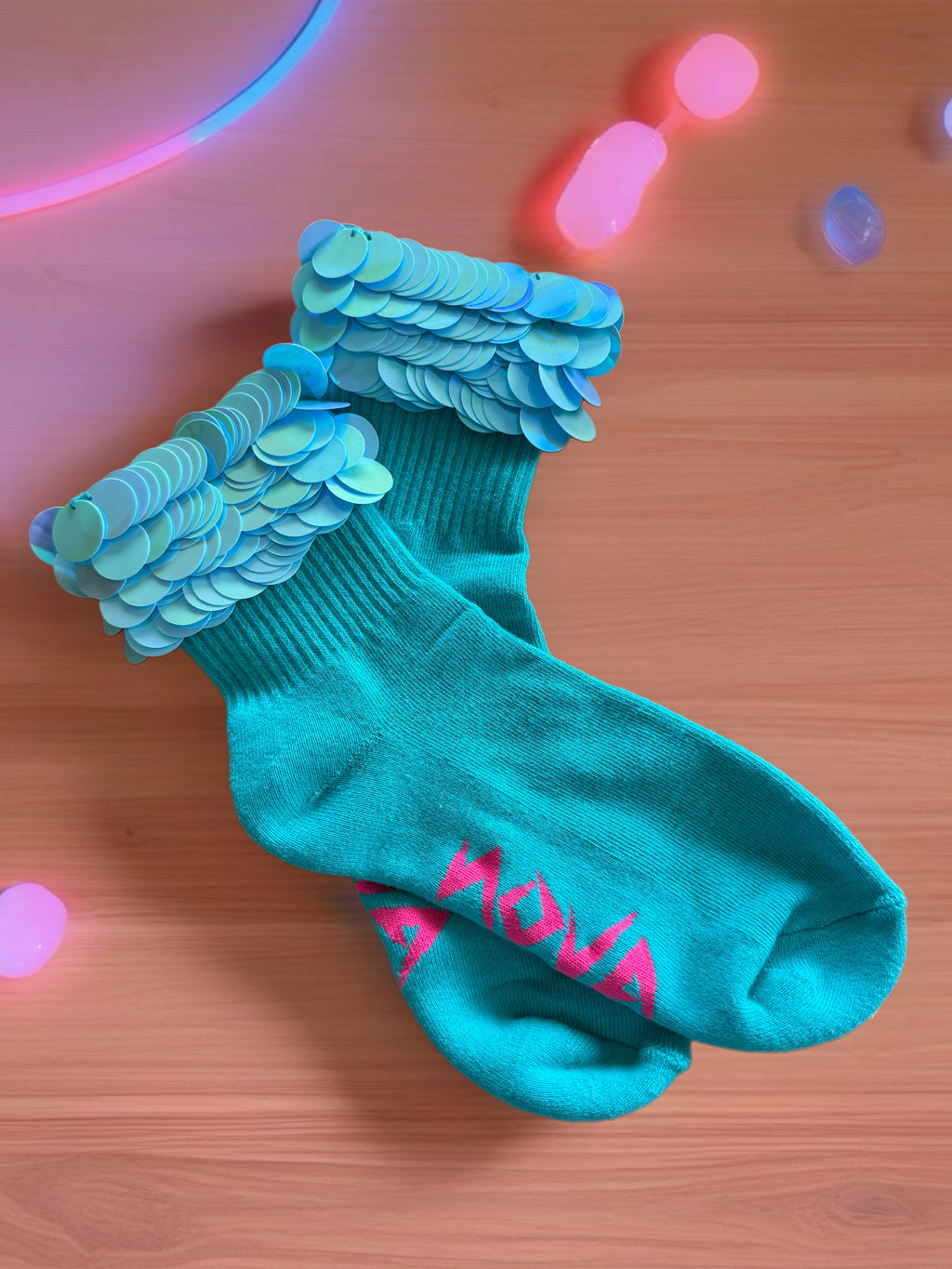 A pair of teal crew socks with light blue sequined cuffs and pink text on the foot stating NOVA brand, displayed on a wooden surface with colorful neon light reflections.